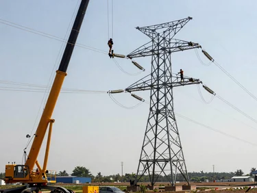 Close-up of high-voltage transmission towers being erected by crane in open terrain, steel lattice arms extended, linemen working aloft, clear sky, wide site visible in background