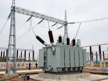 Ground-level view of a high-voltage substation nearing completion, transformer banks installed, cable trays running between steel structures, workers in hard hats visible mid-frame, flat overcast daylight